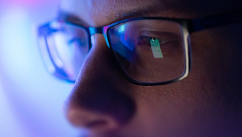 Eine Nahaufnahme einer Person mit einer schwarzen, rechteckigen Brille, auf deren Gläsern sich blaue Bildschirmreflexionen zeigen, während im Hintergrund ein unscharfes Gesicht zu sehen ist.