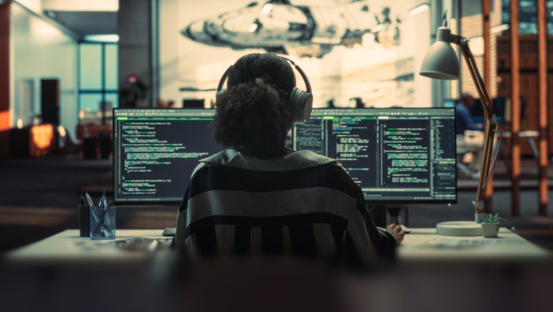 Eine Person mit Kopfhörern sitzt an einem Schreibtisch in einem modernen Büro, vor sich zwei Computerbildschirme, auf denen Code zu sehen ist.